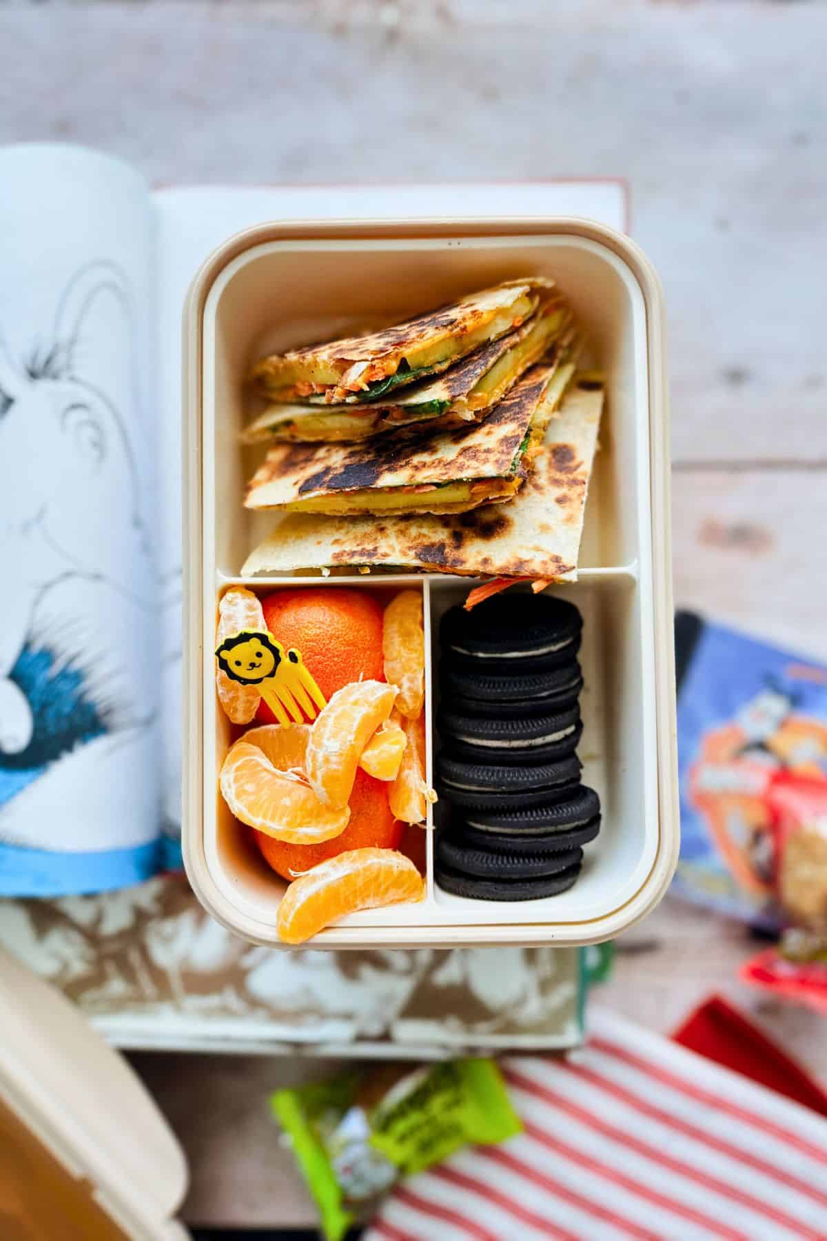 A lunchbox with sliced apple and veggie quesadillas stacked in one compartment, paired with mandarin orange segments and chocolate sandwich cookies in another side compartments. A small yellow lion-shaped food pick is placed among the orange slices. A Dr. Seuss book and snack packaging are visible in the background.