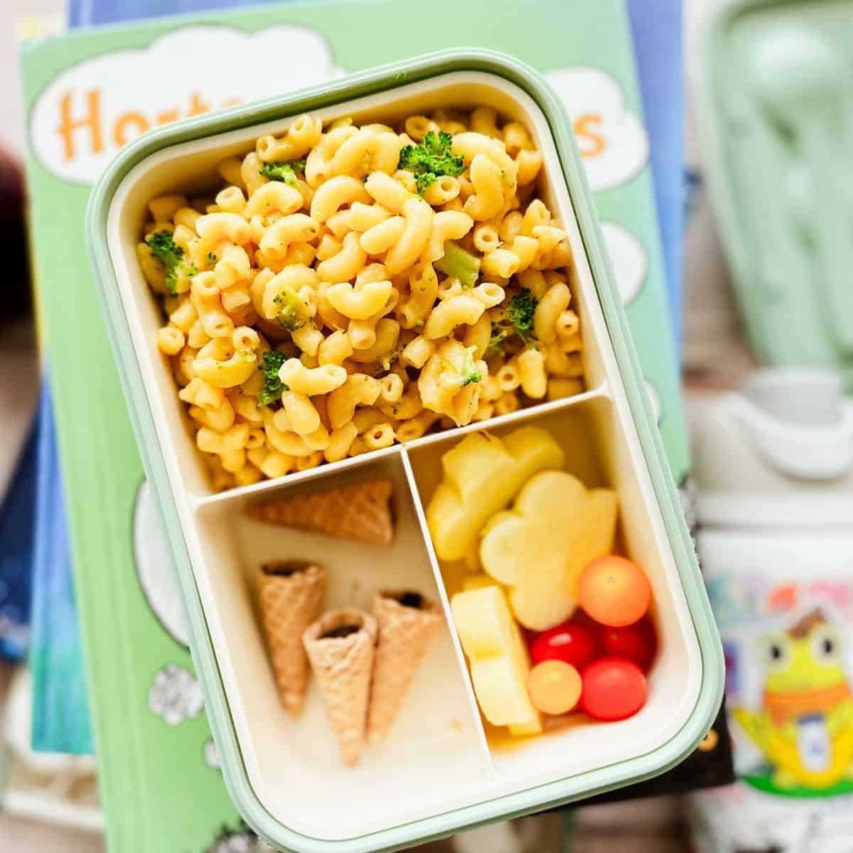 bento box filled with the mac and broccoli in one section with the other sections filled with apples shaped like flowers, small tomatoes, and dessert cone tips.