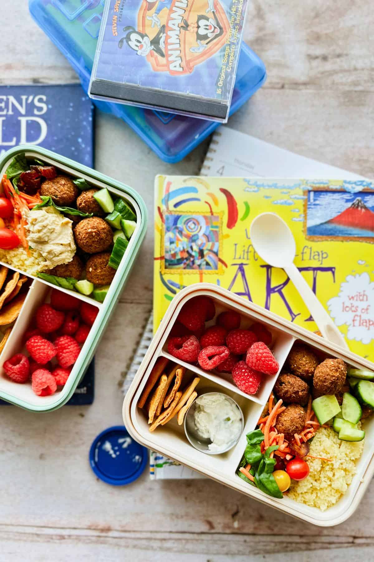 Two bento-style vegan lunchboxes filled with falafel, couscous, cucumbers, cherry tomatoes, shredded carrots, greens, hummus or tzatziki, pita chips, and fresh raspberries. Lunchboxes are placed on top of colorful children’s books and school supplies, creating a vibrant back-to-school setting.