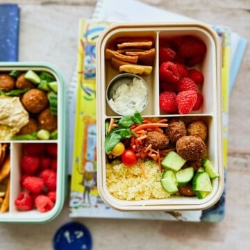Lunchbox containing mini falafel, couscous, chopped cucumber, cherry tomatoes, shredded carrots, spinach, pita chips, a small container of dairy-free tzatziki, and fresh raspberries. The lunchbox is placed on top of children’s books and notebooks, styled for a back-to-school theme.