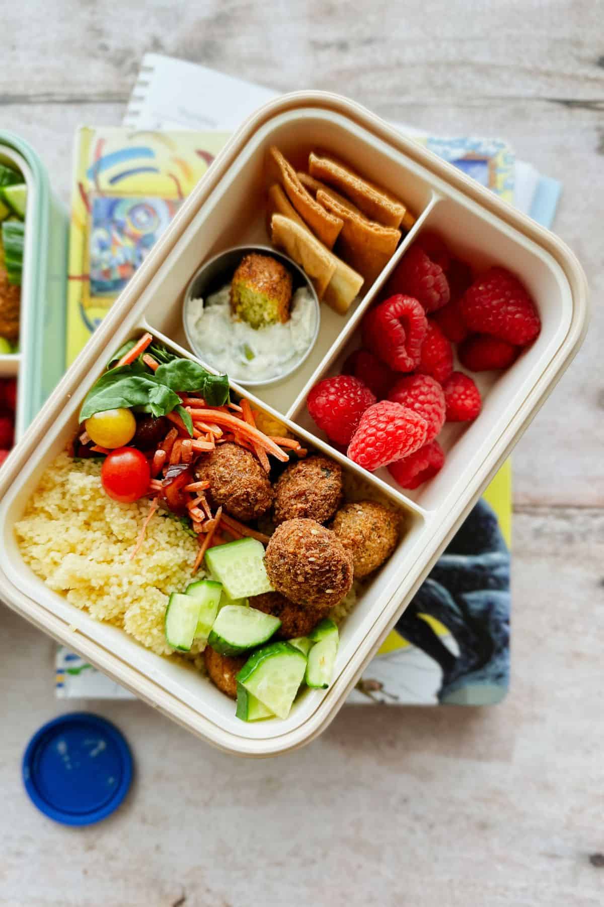 Bento-style vegan lunchbox featuring mini falafel, couscous, chopped cucumber, shredded carrots, spinach, cherry tomatoes, raspberries, pita chips, and a small container of vegan tzatziki with a falafel dipped inside. Lunchbox is placed on top of colorful children’s books, styled for a back-to-school setting.