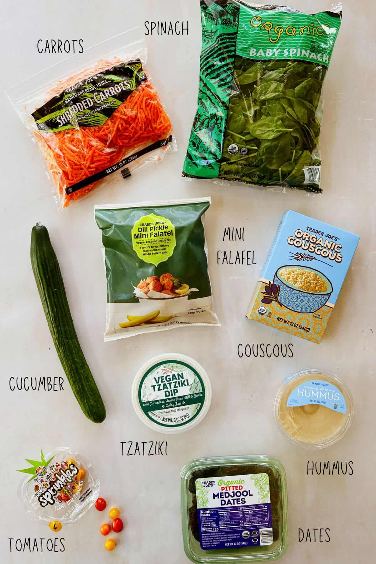 Flat lay of ingredients for a vegan Mediterranean lunch bowl, including Trader Joe’s shredded carrots, organic baby spinach, dill pickle mini falafel, organic couscous, vegan tzatziki dip, hummus, English cucumber, cherry tomatoes, and organic pitted Medjool dates. Each item is labeled with text on a white background.