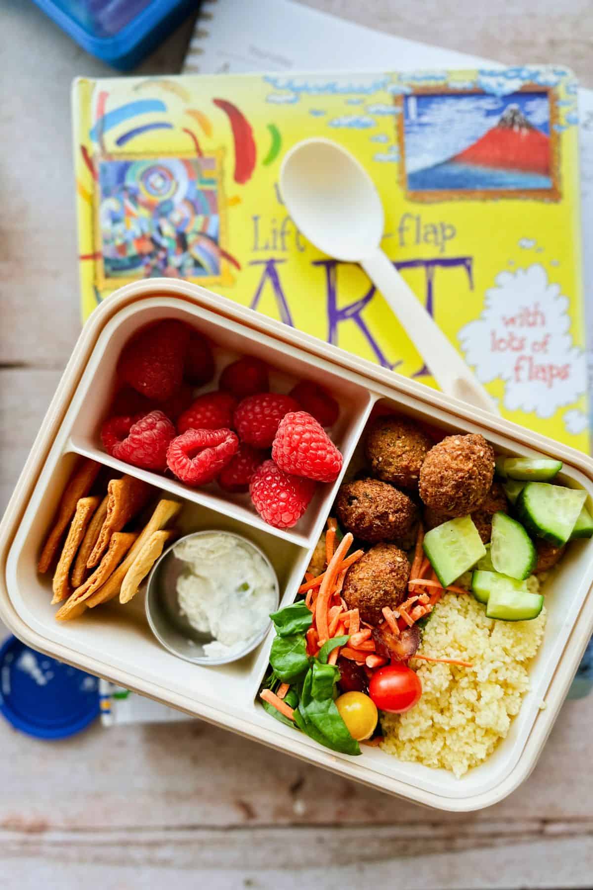 Bento-style vegan lunchbox with falafel, couscous, cucumber, shredded carrots, cherry tomatoes, spinach, pita chips, a small container of tzatziki, and fresh raspberries. Lunchbox is placed on top of a colorful children’s art book with a spoon beside it.