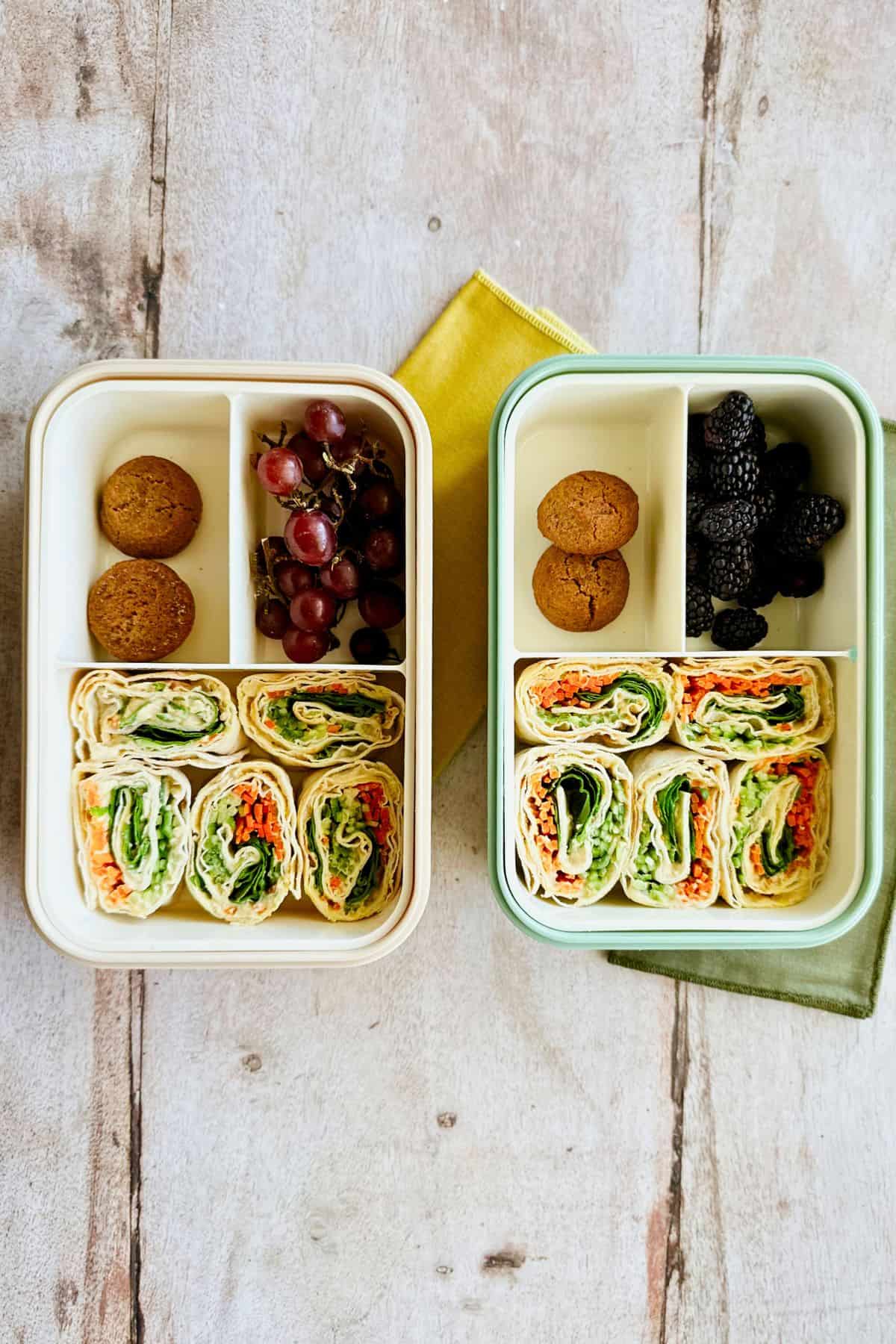 Two bento boxes with the hummus and veggie pinwheels, cookies, blackberries, and grapes.
