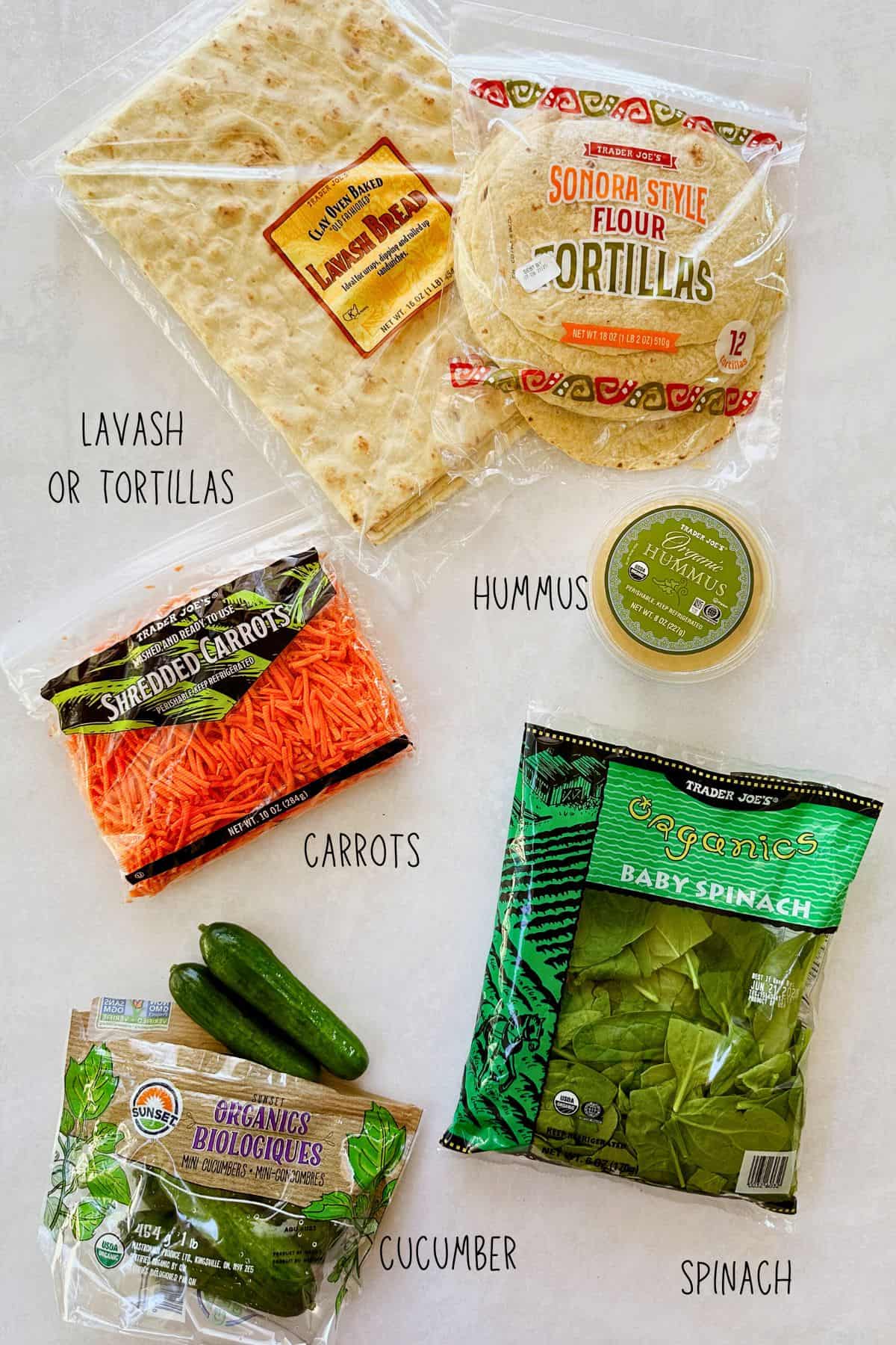 ingredients for pinwheels layed on table. They include shredded carrots, cucumbers, spinach, hummus, lavash, and tortillas.