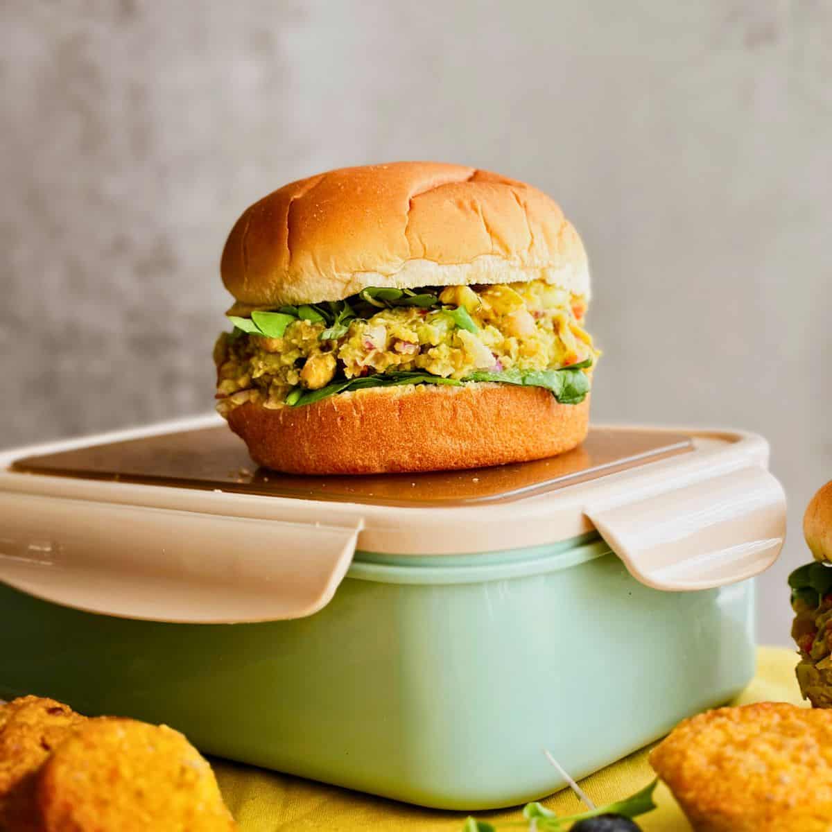 A close-up of a fluffy sandwich bun filled with creamy no-mayo chickpea salad and leafy greens, resting on top of a mint green lunch container surrounded by mini cornbread muffins.