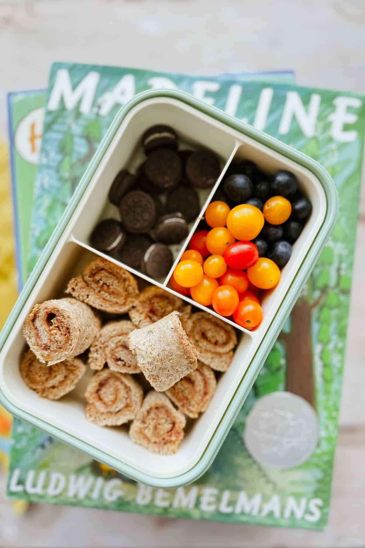 Nut and Butter Jelly Rolls in a bento box along with mini oreos, blueberries, and mini tomatoes. The bento box is stacked on children's books.