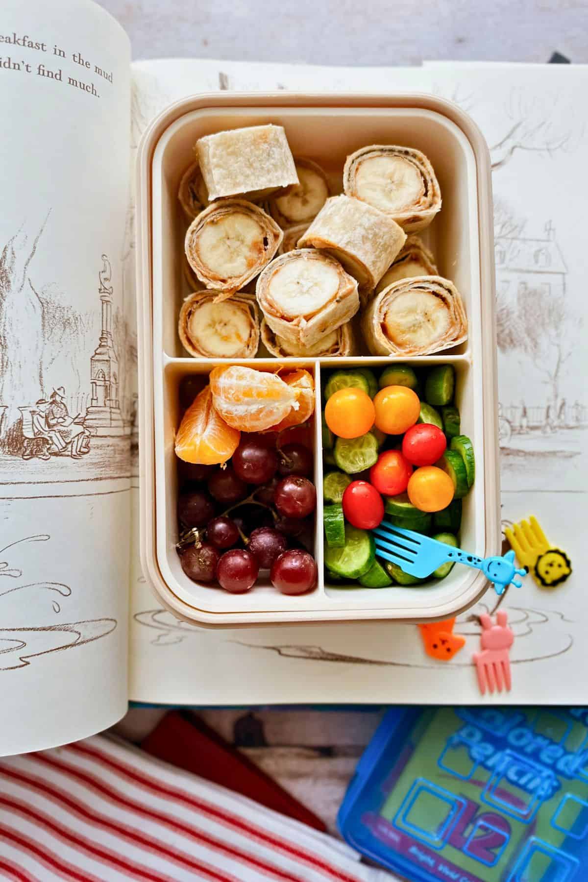 A bento-style lunchbox filled with peanut butter and banana roll-ups made from whole wheat tortillas, alongside red grapes, orange slices, mini cucumbers, and colorful cherry tomatoes with a blue giraffe fork. The lunchbox is set on top of an open illustrated children’s book.