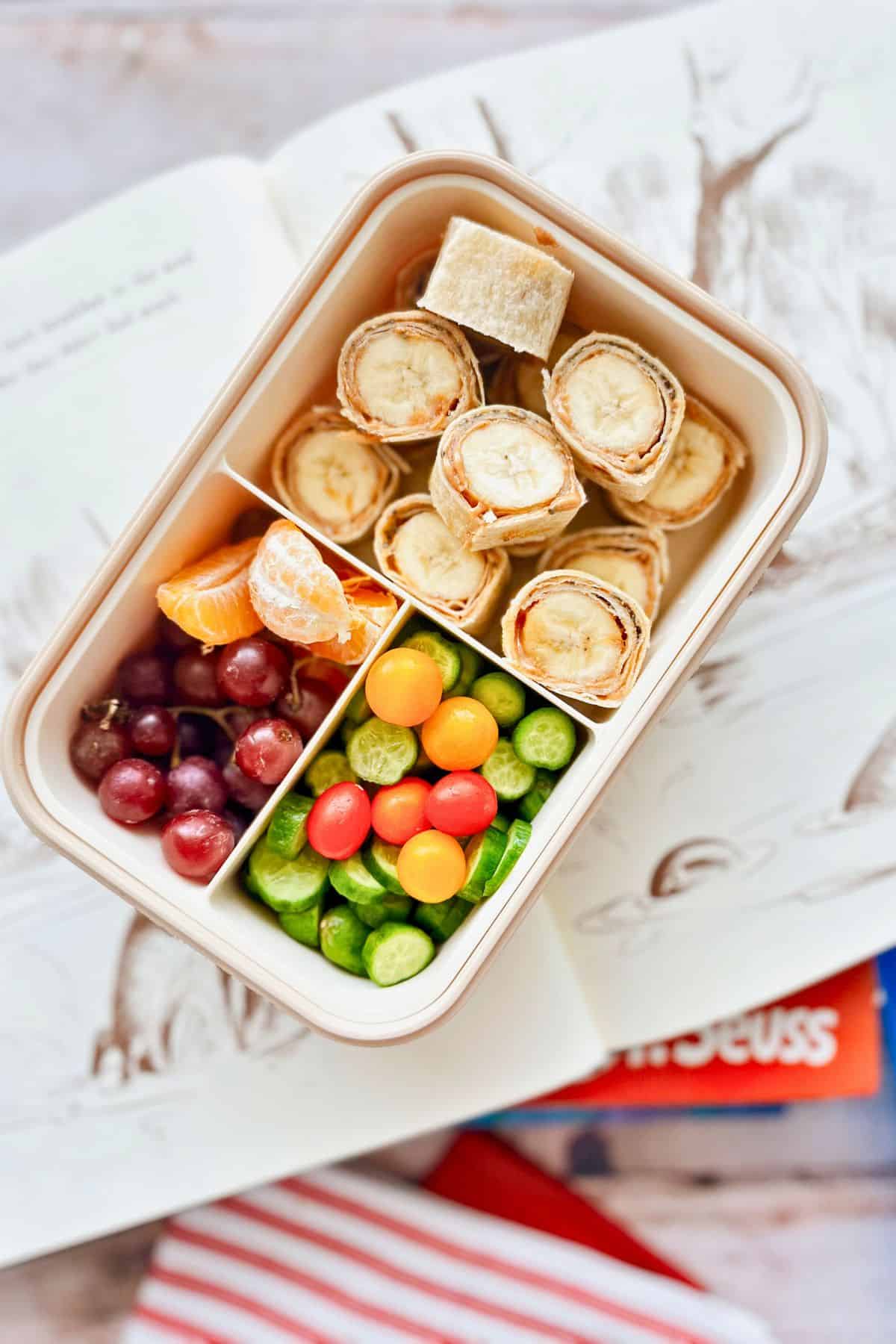 A kid’s bento-style lunchbox featuring peanut butter and banana roll-ups, red grapes, mandarin orange slices, halved cherry tomatoes, and sliced cucumbers. The lunchbox is placed on top of an open illustrated children’s book with a red-and-white striped napkin and a Dr. Seuss book visible underneath.
