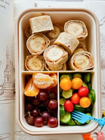 A bento-style lunchbox filled with peanut butter and banana roll-ups made from whole wheat tortillas, alongside red grapes, orange slices, mini cucumbers, and colorful cherry tomatoes with a blue giraffe fork. The lunchbox is set on top of an open illustrated children’s book.