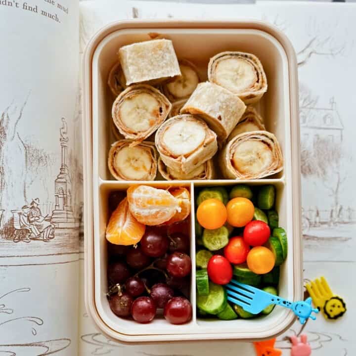A bento-style lunchbox filled with peanut butter and banana roll-ups made from whole wheat tortillas, alongside red grapes, orange slices, mini cucumbers, and colorful cherry tomatoes with a blue giraffe fork. The lunchbox is set on top of an open illustrated children’s book.