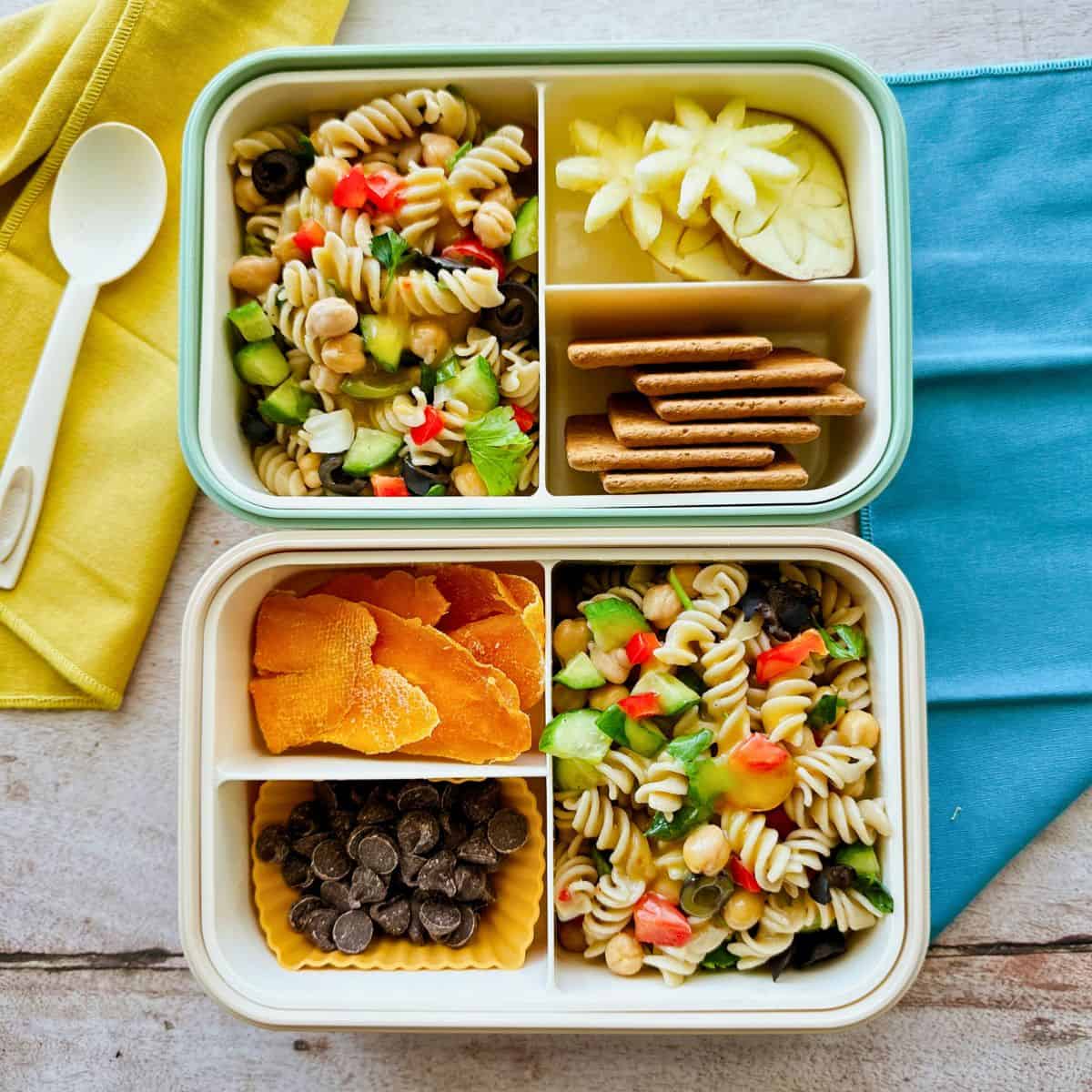 two bento boxes with the quinoa pasta salad served with sides such as graham crackers, apples cut into flowers, dairy free chocolate chips, and dried mango.