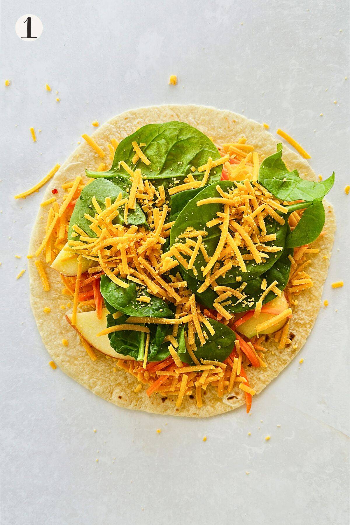 A tortilla topped with shredded carrots, fresh spinach leaves, apple slices, and vegan cheddar cheese, ready to be topped with another tortilla.
