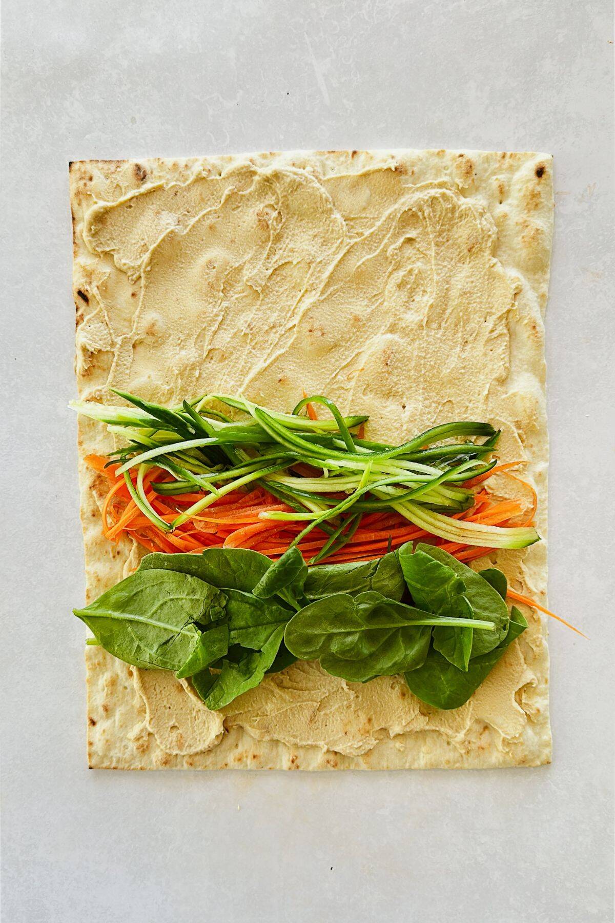 lavash on a table with hummus spread on it and veggies placed in rows before rolling.