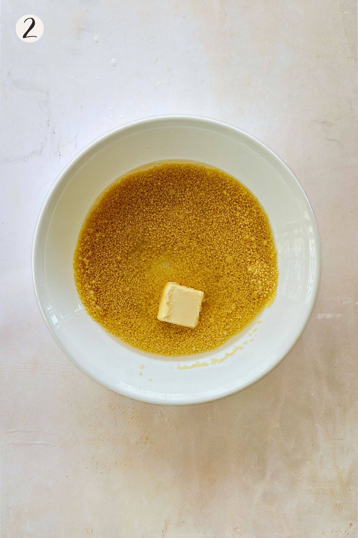 Dry couscous soaking in a white bowl with a cube of plant-based butter on top, ready for steaming.