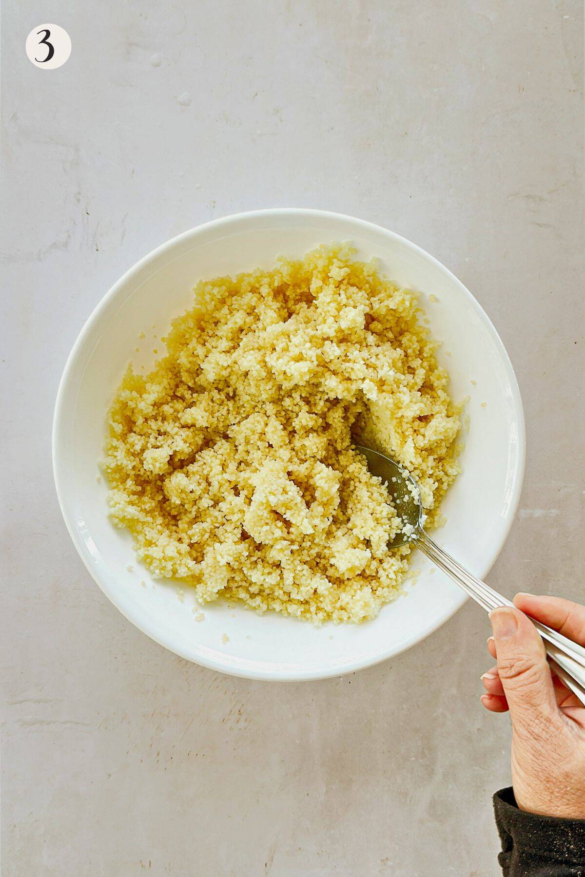 Cooked couscous fluffed with a spoon in a white bowl, ready to be used as a base for a lunch bowl.