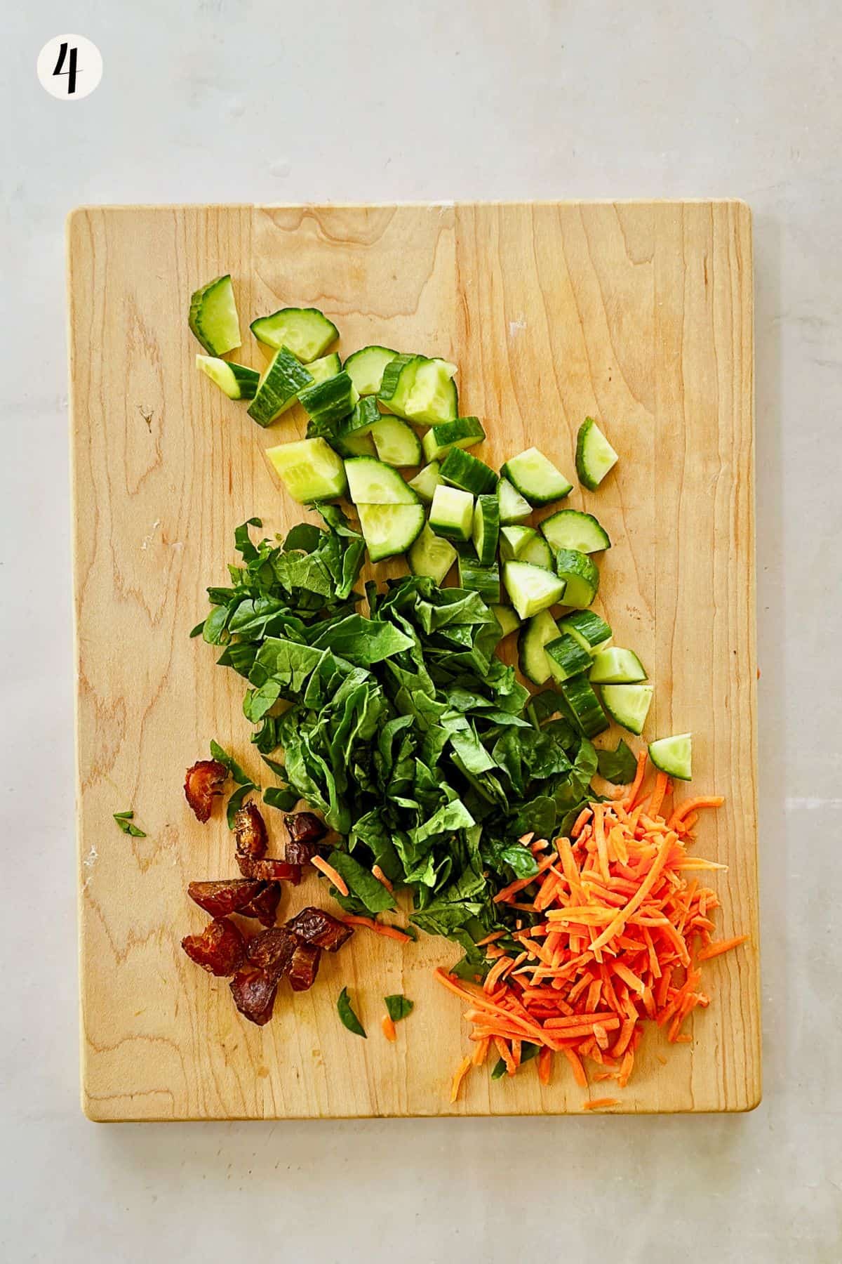 Chopped cucumber, shredded carrots, baby spinach, and diced Medjool dates arranged on a wooden cutting board for a vegan Mediterranean lunch bowl.