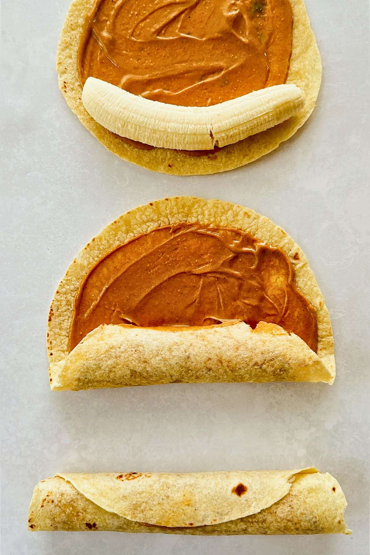 Step-by-step visual of making peanut butter and banana roll-ups—three tortillas shown in stages: one topped with peanut butter and a banana, one partially rolled, and one fully rolled on a light surface.
