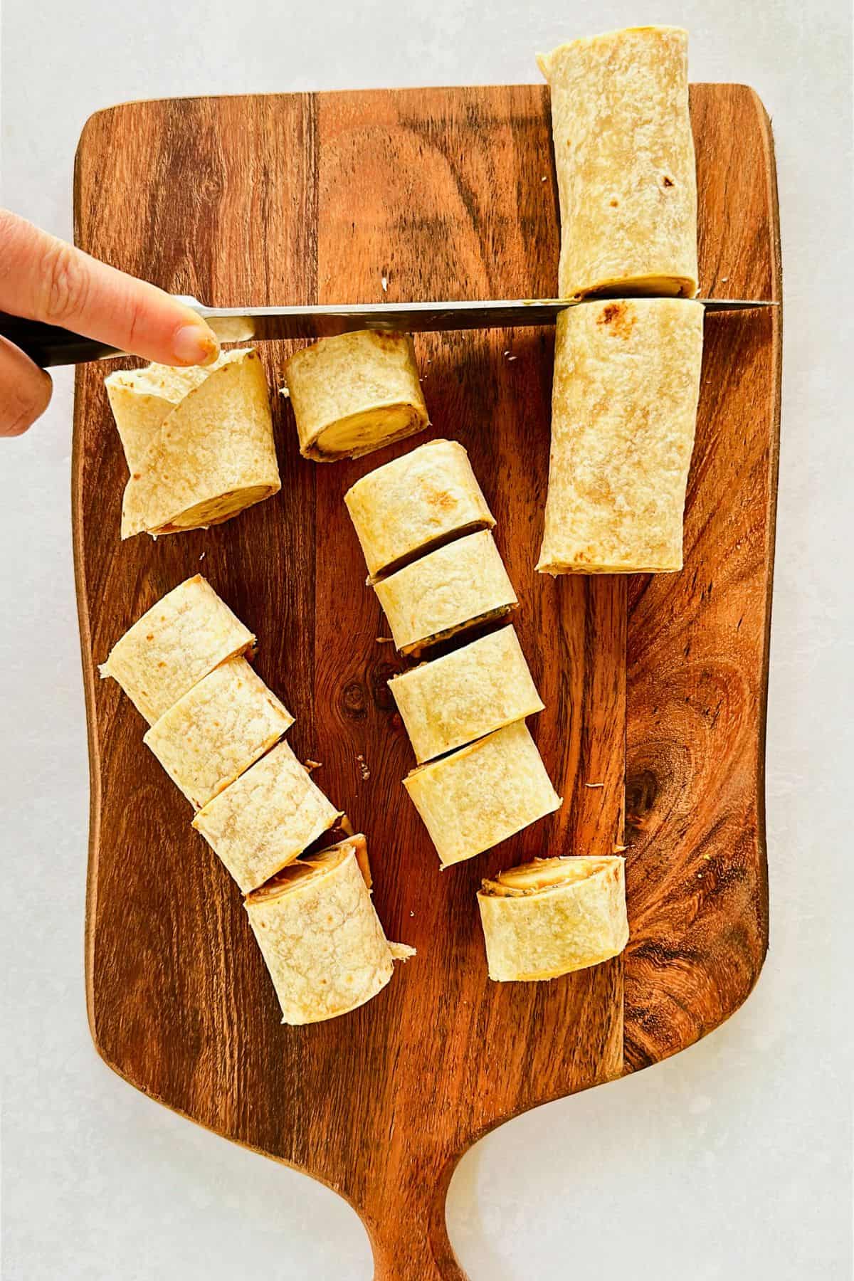 A hand slicing peanut butter and banana roll-ups into bite-sized pieces on a wooden cutting board. Rolled tortillas are neatly cut into pinwheels.