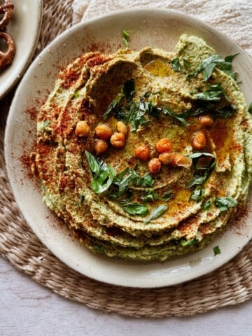 Plate of creamy basil hummus topped with crispy chickpeas, paprika, and fresh herbs, served on a woven placemat.