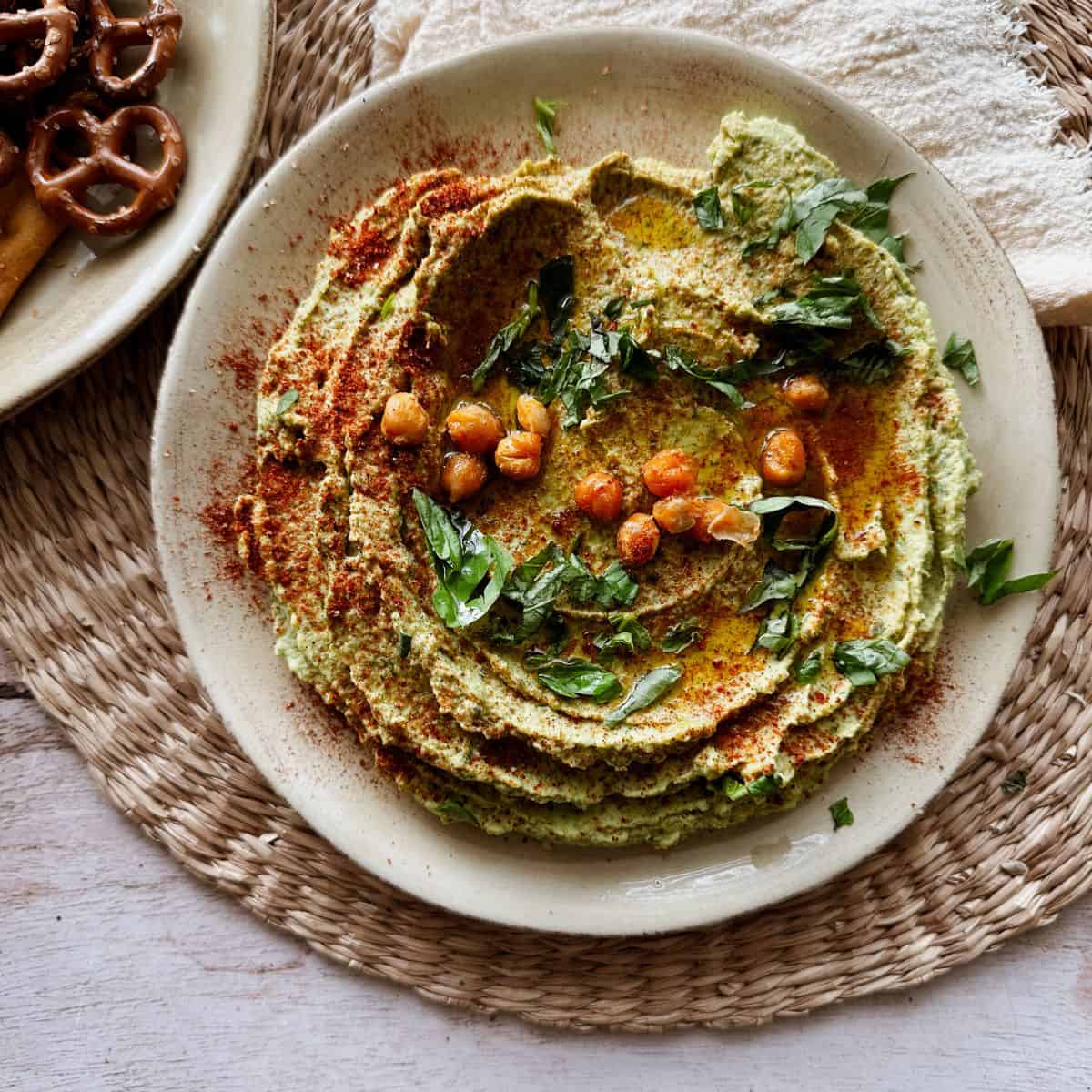 Plate of creamy basil hummus topped with crispy chickpeas, paprika, and fresh herbs, served on a woven placemat.