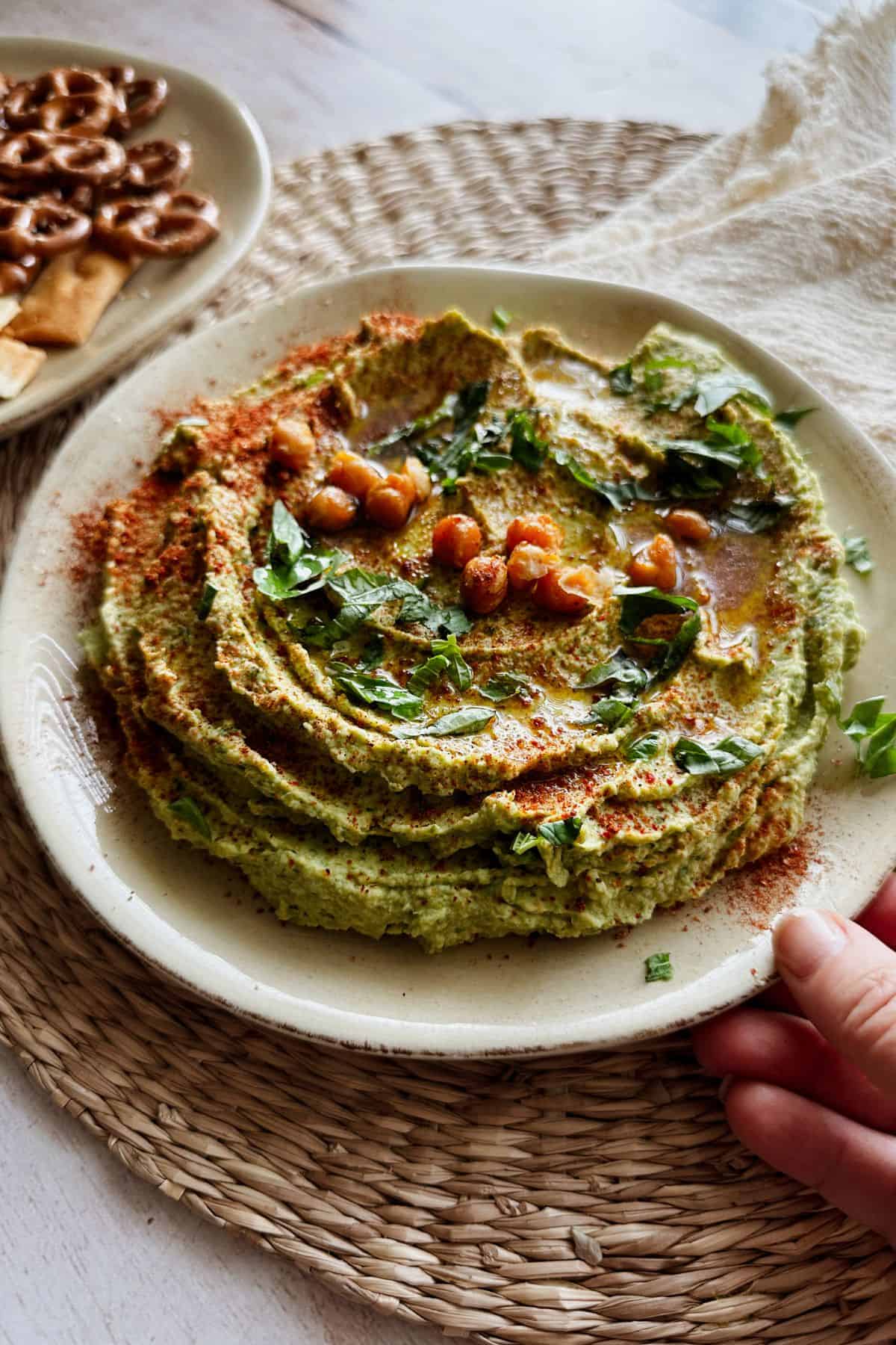 Hand holding a plate of creamy basil hummus topped with crispy chickpeas, fresh herbs, paprika, and olive oil, served on a woven placemat with pretzels and pita chips in the background.