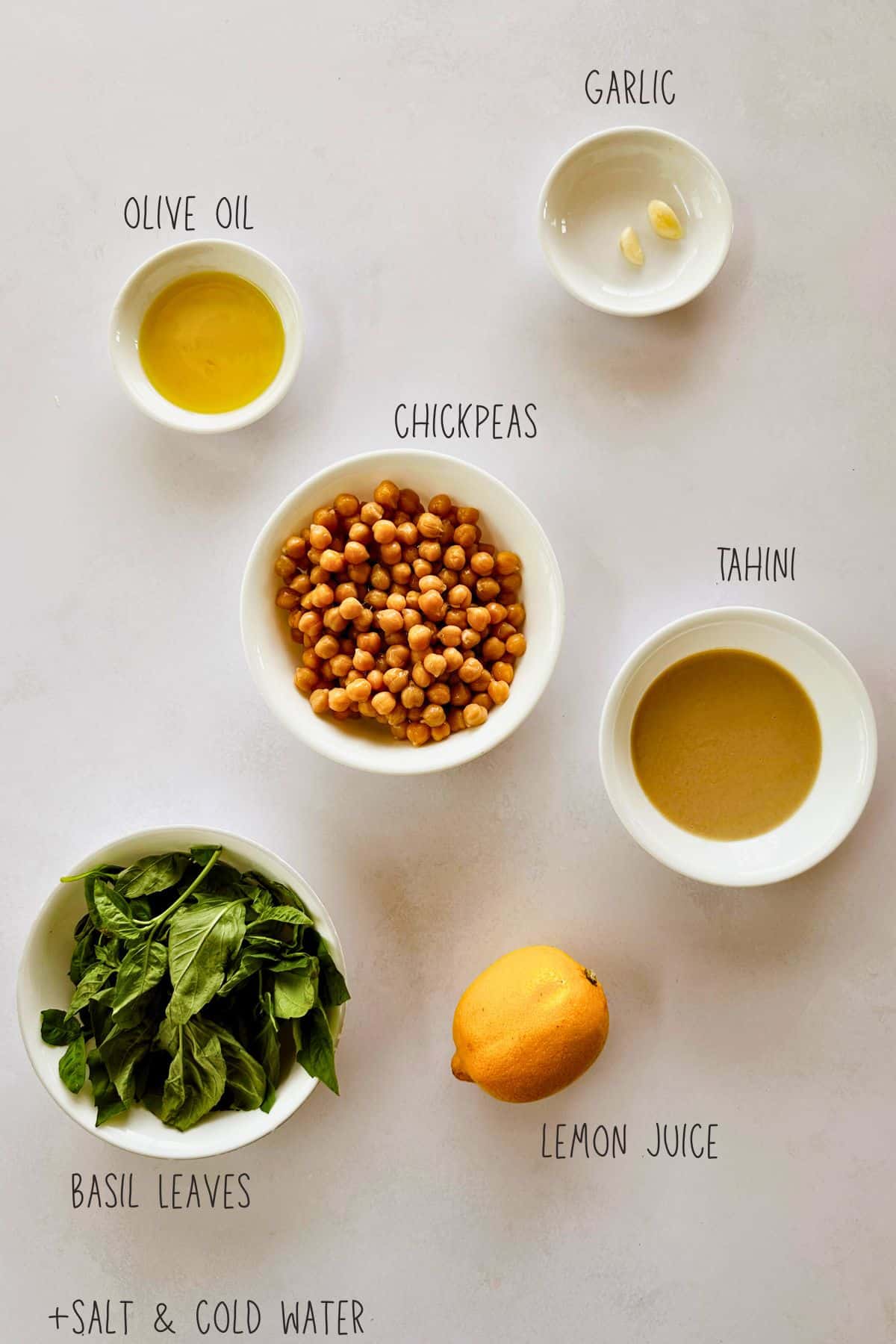 Flat lay of labeled ingredients for basil hummus, including olive oil, garlic, chickpeas, tahini, basil leaves, lemon juice, salt, and cold water. All items are arranged in small white bowls on a light-colored background.