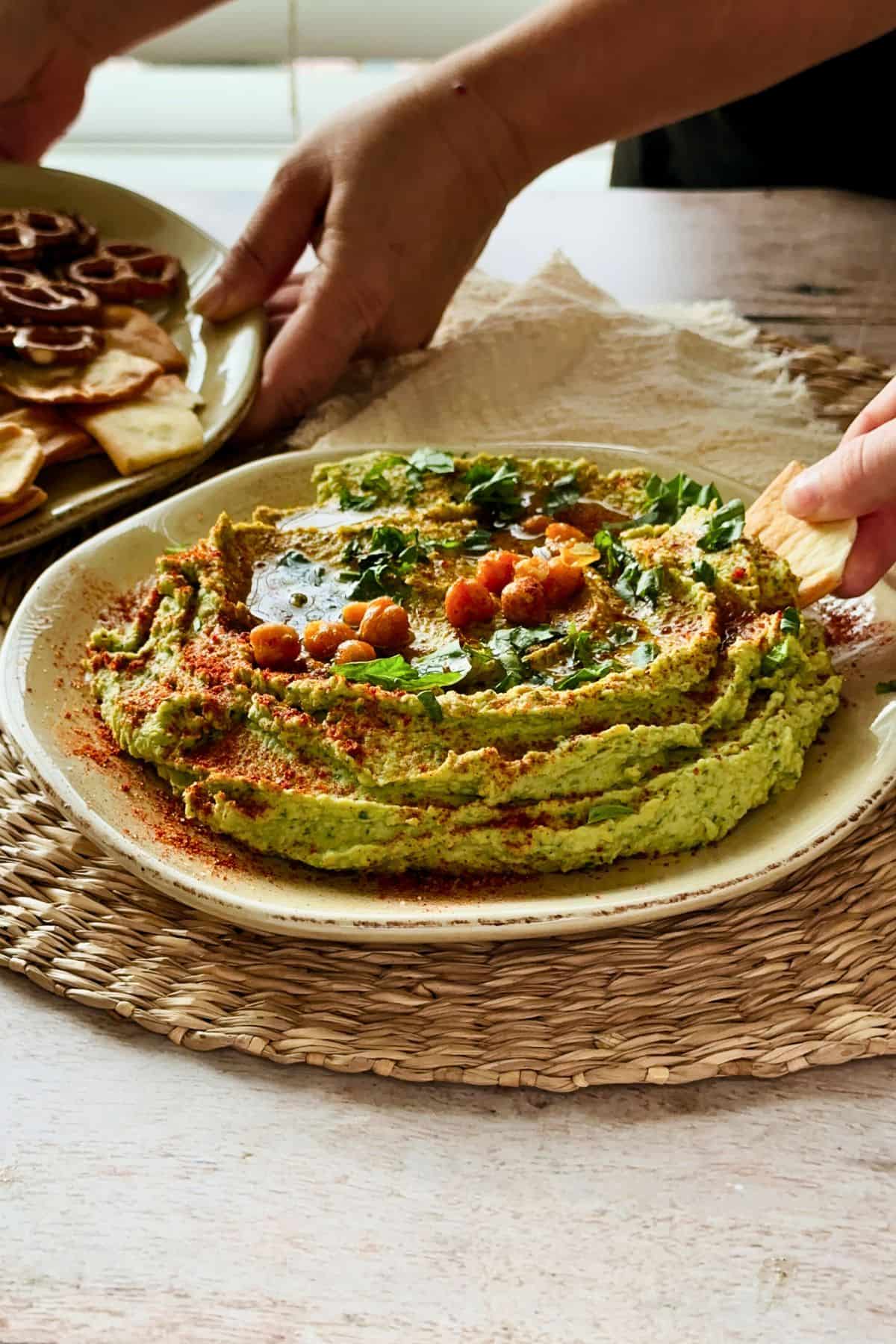 Hands serving a plate of pretzels and crackers alongside a bowl of green basil hummus topped with olive oil, chickpeas, and basil, with another hand dipping a cracker.