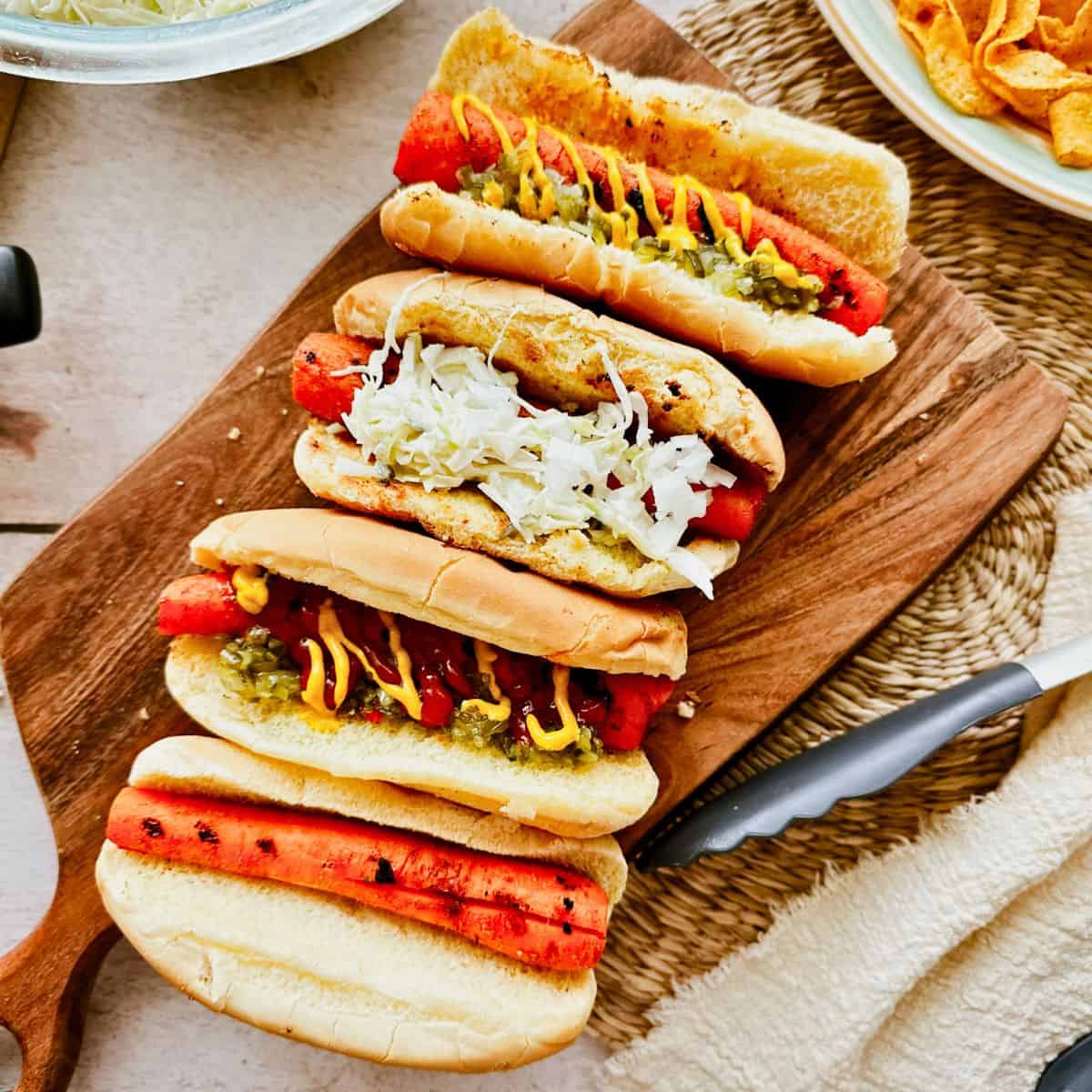 Overhead view of four grilled carrot dogs in toasted buns, arranged on a wooden board. Each one is topped differently-with mustard and relish, shredded cabbage, ketchup, and one served plain-highlighting a variety of topping ideas for vegan carrot hot dogs. A bowl of vegan coleslaw and a plate of chips are partially visible in the background.