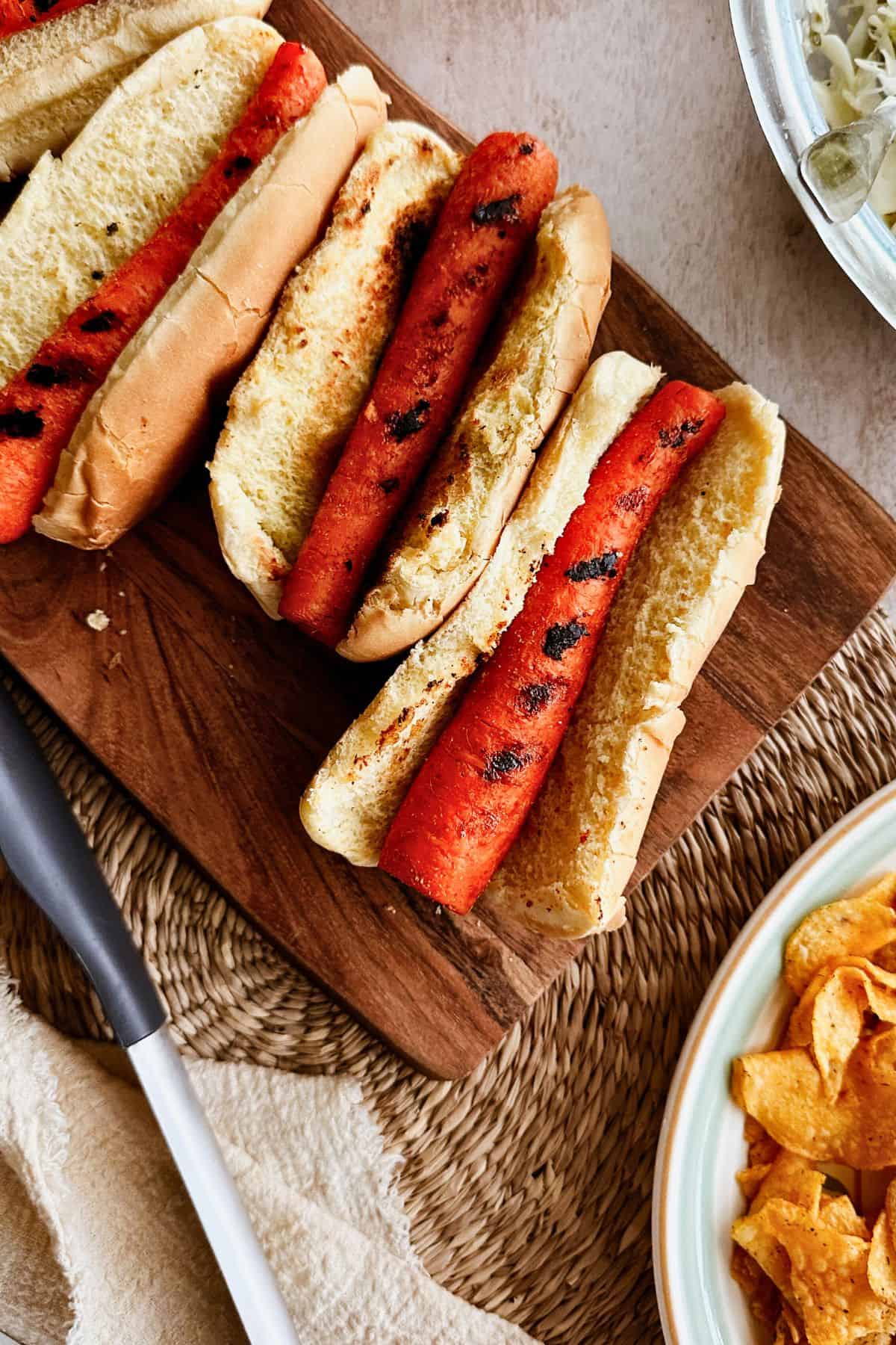 A close-up view of grilled carrot dogs nestled in toasted hot dog buns, resting on a wooden cutting board. The carrots have visible grill marks, and a bowl of potato chips is partially visible on the side.