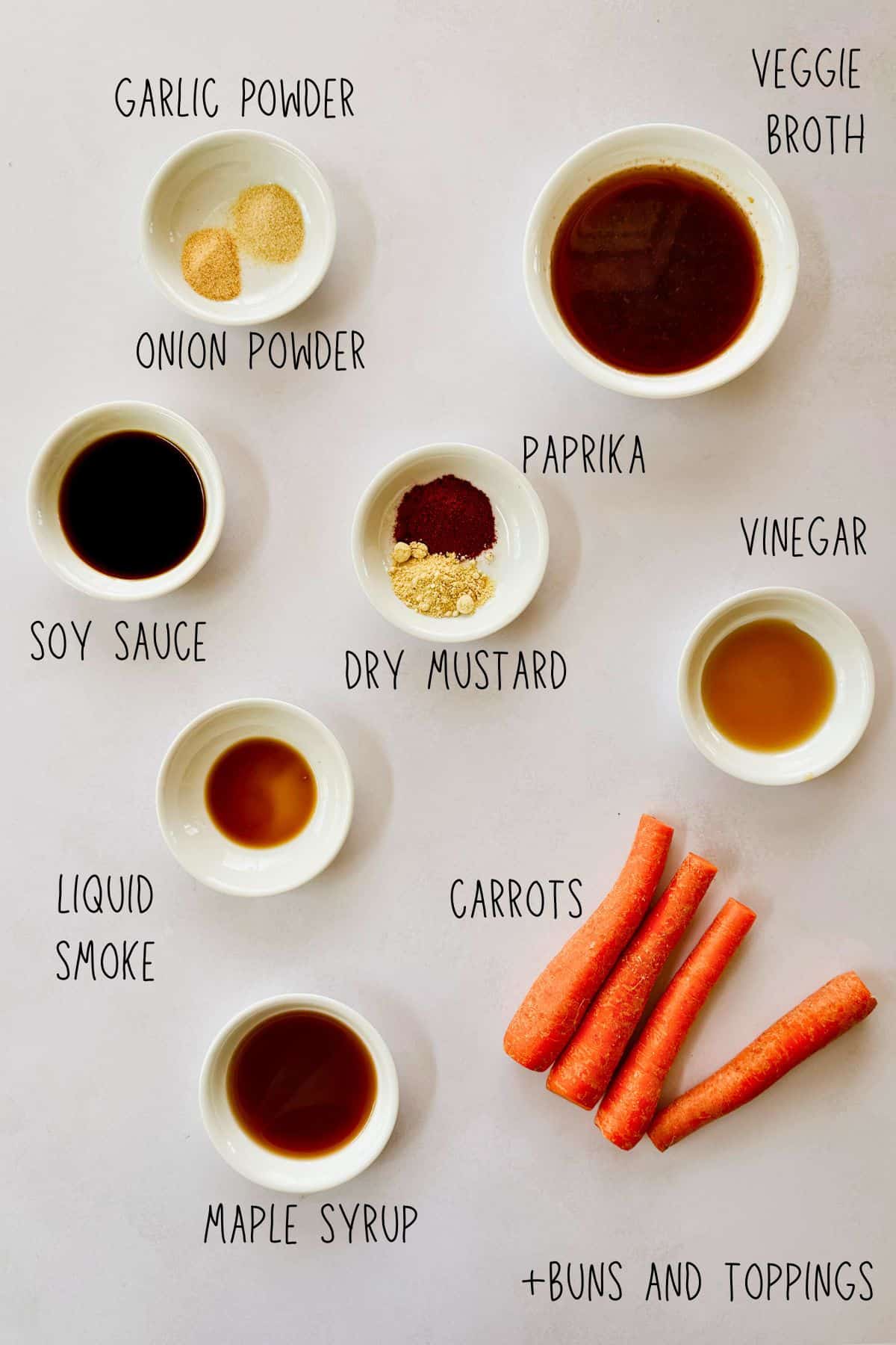 Overhead image showing labeled ingredients for making vegan carrot dogs. Includes garlic powder, onion powder, soy sauce, liquid smoke, maple syrup, paprika, dry mustard, vinegar, veggie broth, whole carrots, and a note for buns and toppings. Ingredients are arranged in small white bowls on a light surface.