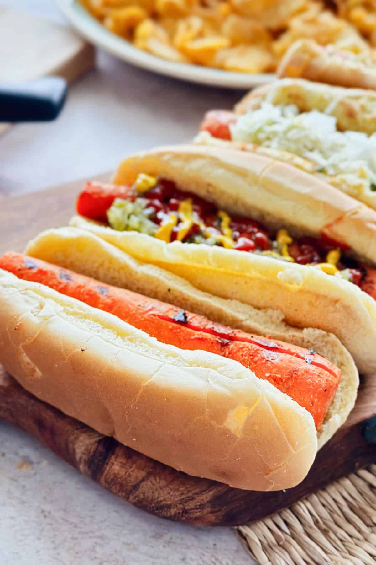 Close-up of a grilled carrot dog in a soft hot dog bun, showing char marks and a juicy texture, with other topped carrot dogs slightly blurred in the background and a plate of chips in the distance.