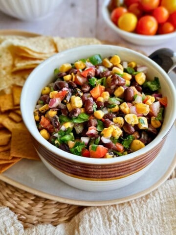A vibrant bowl of Corn and Black Bean Salsa made with black beans, roasted corn, red onion, diced tomatoes, and fresh herbs. The bowl is surrounded by tortilla chips and crackers on a woven placemat, ready to serve as a dip or side dish. Bright cherry tomatoes are blurred in the background.