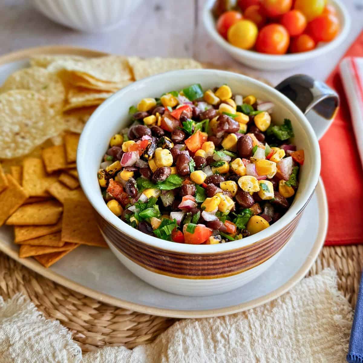 A vibrant bowl of Corn and Black Bean Salsa made with black beans, roasted corn, red onion, diced tomatoes, and fresh herbs. The bowl is surrounded by tortilla chips and crackers on a woven placemat, ready to serve as a dip or side dish. Bright cherry tomatoes are blurred in the background.