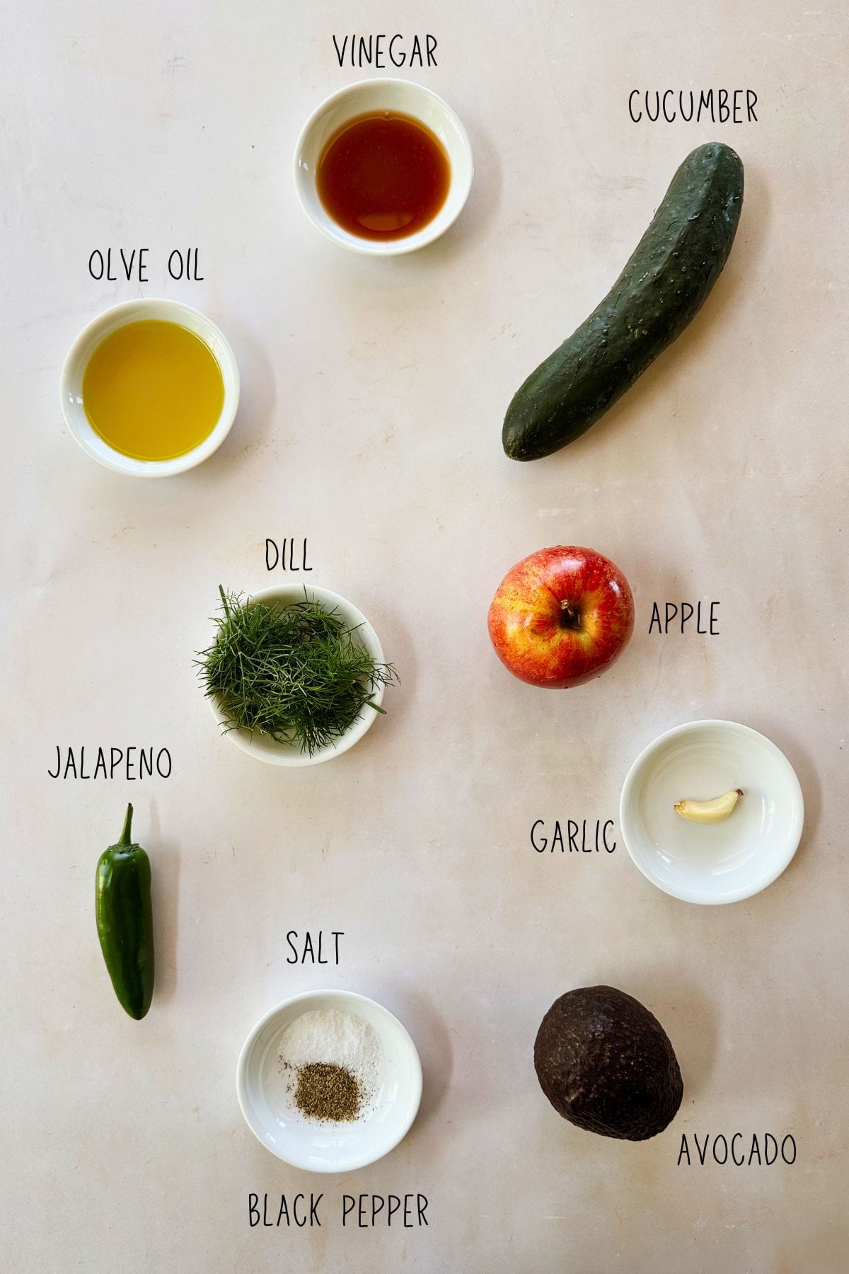 Labeled overhead photo of ingredients for cucumber gazpacho, including olive oil, vinegar, cucumber, apple, avocado, garlic, jalapeño, dill, salt, and black pepper in small bowls arranged on a light surface.