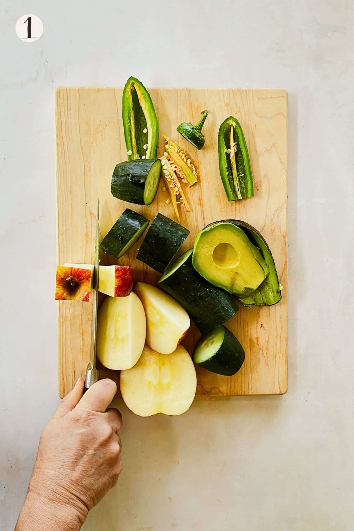 Overhead view of chopped cucumber, apple, avocado, and jalapeño on a wooden cutting board. A hand slices an apple while prepping ingredients for cucumber gazpacho.