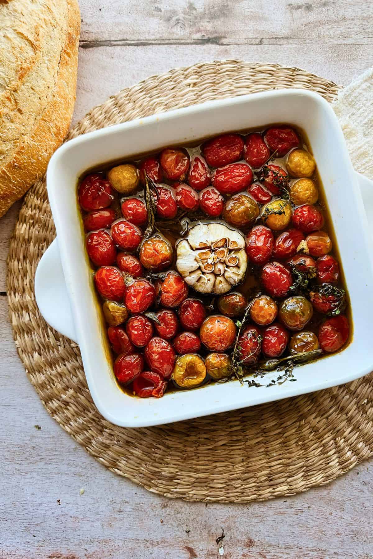A white ceramic baking dish filled with slow-roasted cherry tomatoes and a cut head of garlic, glistening in olive oil with sprigs of herbs. The dish rests on a woven placemat, next to a crusty loaf of bread.