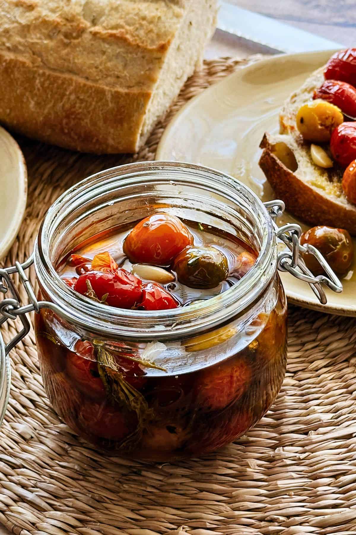 A glass jar filled with vibrant cherry tomato confit, swimming in olive oil with garlic and herbs, set on a woven placemat next to crusty bread.