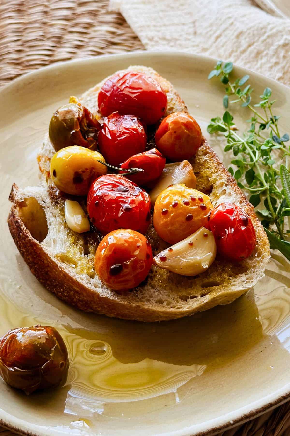 A slice of rustic bread topped with blistered cherry tomatoes, golden garlic, and a drizzle of tomato-infused oil, garnished with fresh herbs.