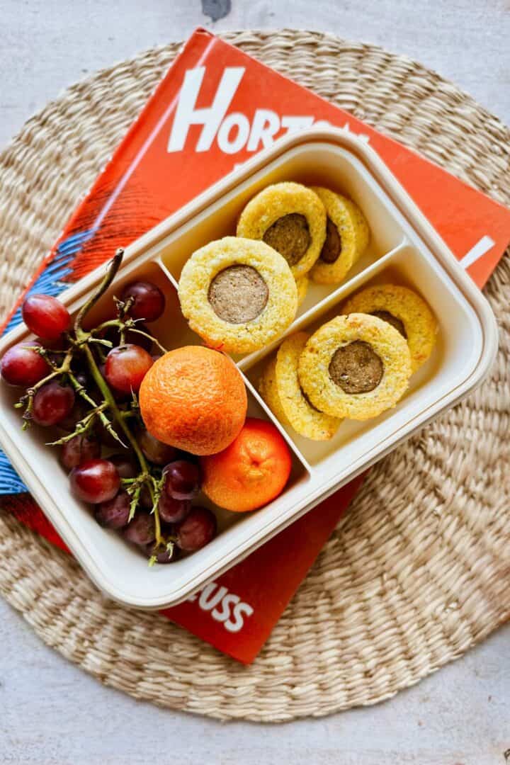 A bento-style lunchbox filled with vegan corn dog bites, a bunch of red grapes, and two small oranges, placed on a woven mat with a Dr. Seuss book in the background.