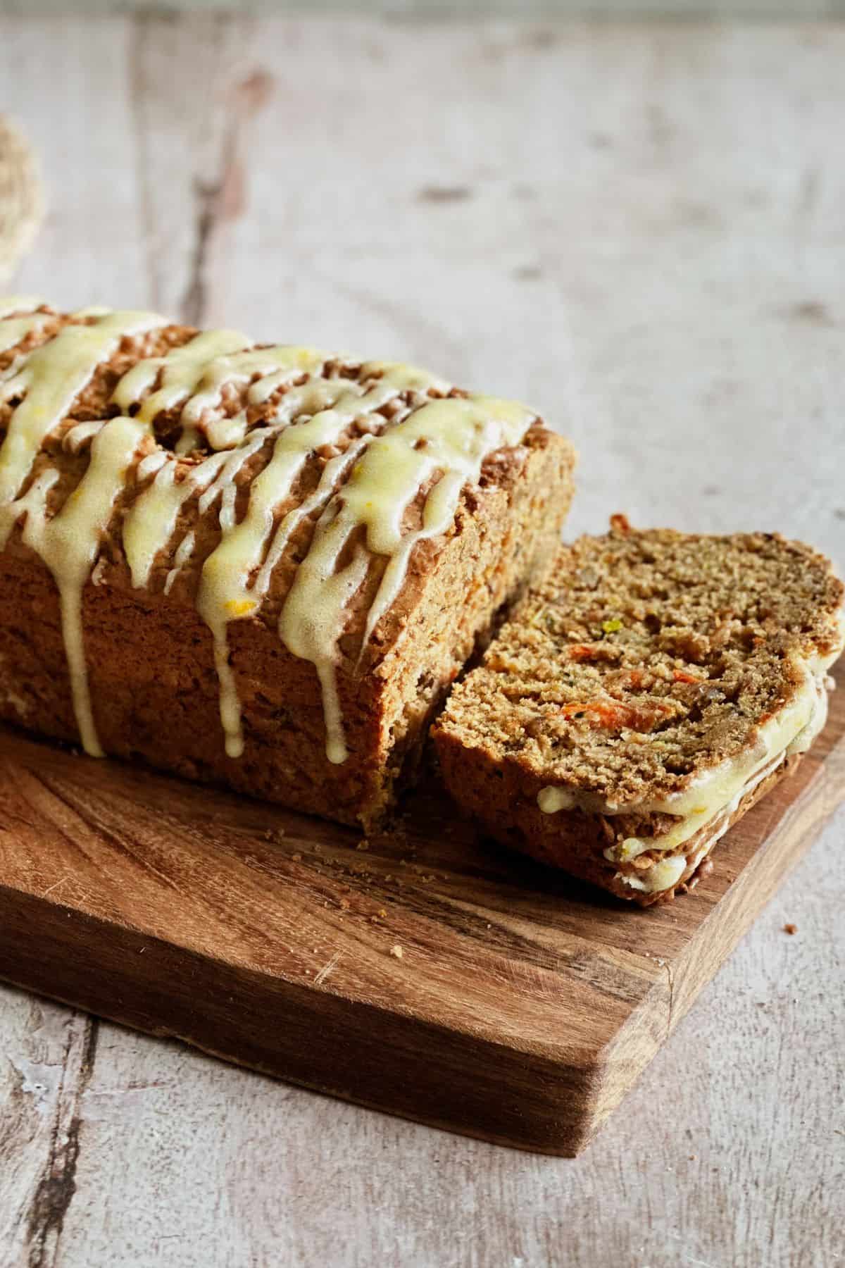 Close-up of a sliced loaf of zucchini, carrot, and apple bread topped with a drizzle of orange cream cheese glaze, displayed on a wooden cutting board. The texture of the moist bread and visible veggie shreds are highlighted.