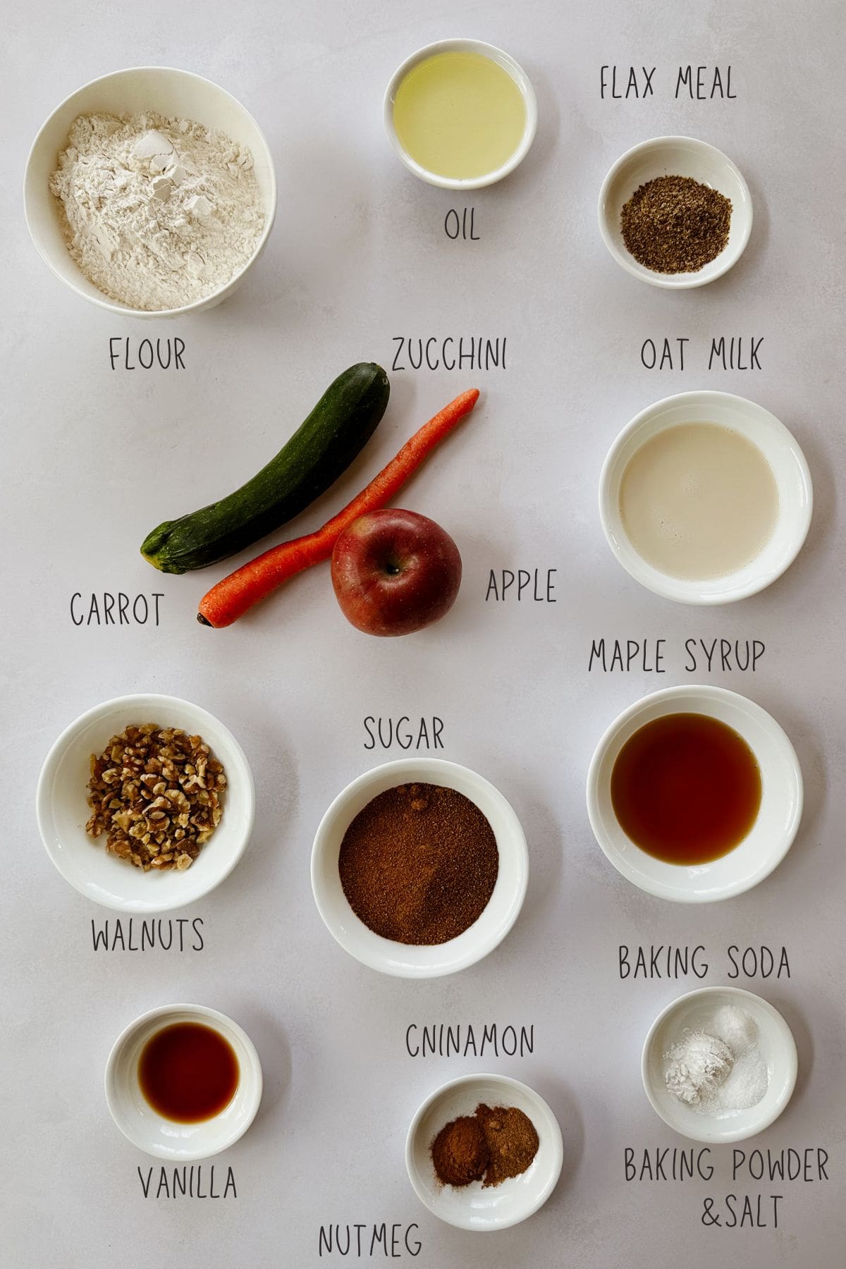 Flat lay of labeled ingredients for vegan carrot apple zucchini bread, arranged on a light surface. Includes flour, flax meal, oil, oat milk, zucchini, carrot, apple, sugar, maple syrup, baking soda, baking powder, salt, cinnamon, nutmeg, vanilla, and chopped walnuts. Ingredients are placed in small white bowls or as whole produce.