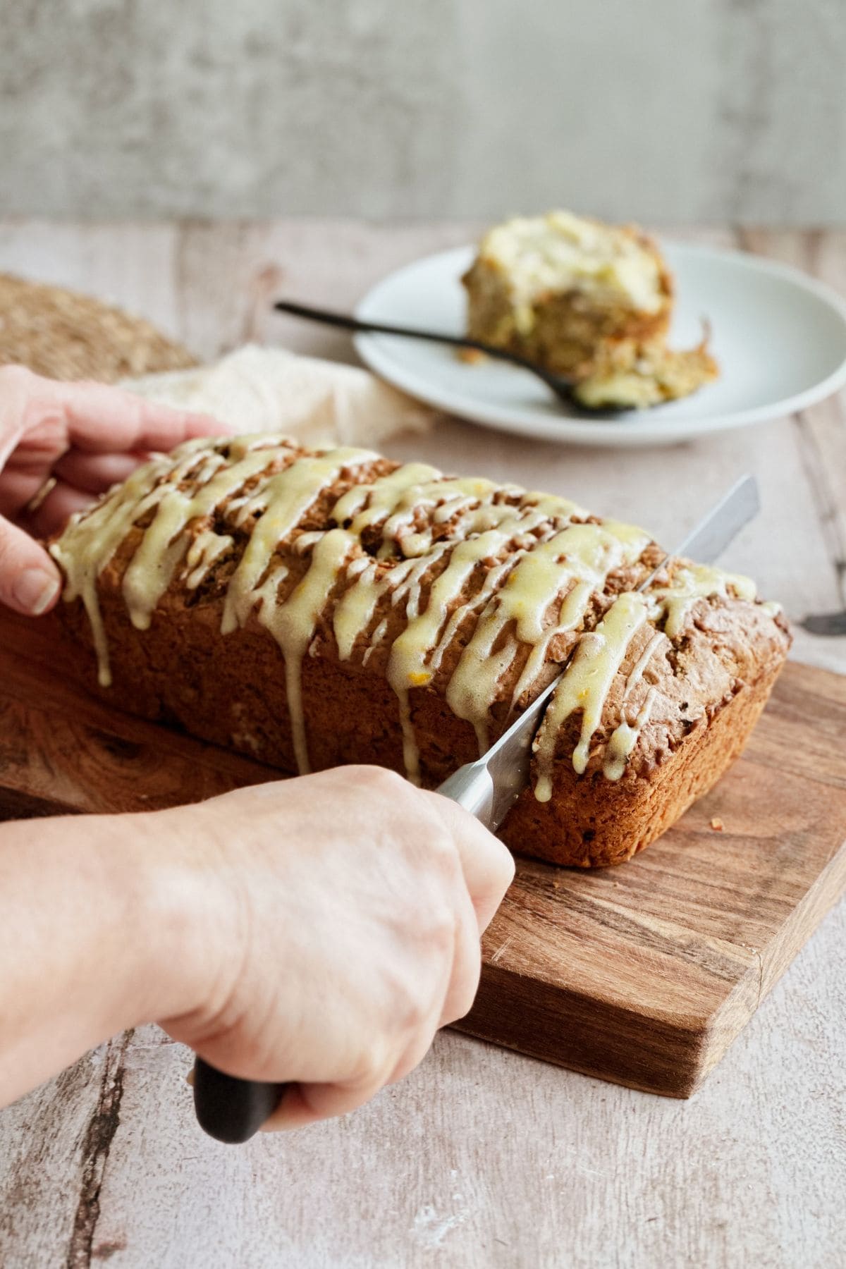 Close-up of hands slicing a loaf of carrot apple and zucchini bread topped with orange cream cheese glaze, set on a wooden cutting board. A slice is served on a plate in the background with a fork.