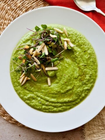 Overhead shot of creamy cucumber gazpacho in a white bowl, garnished with microgreens and toasted slivered almonds. A red napkin and silver spoon rest beside it, with a second bowl of soup partially visible in the corner.