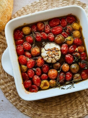 A white ceramic baking dish filled with slow-roasted cherry tomatoes and a cut head of garlic, glistening in olive oil with sprigs of herbs. The dish rests on a woven placemat, next to a crusty loaf of bread.