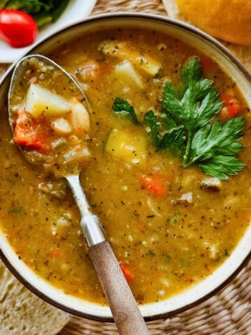 A rustic bowl of zucchini and potato soup with chunks of vegetables and white beans, topped with fresh parsley and a spoon resting in the bowl, ready for a bite. A slice of crusty bread and a salad peek into the frame.