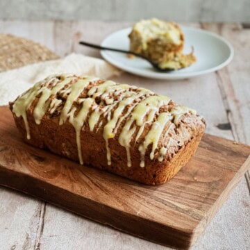 Loaf of zucchini, carrot, and apple bread drizzled with orange cream cheese glaze, resting on a wooden cutting board. A slice is served on a plate in the background with a fork.
