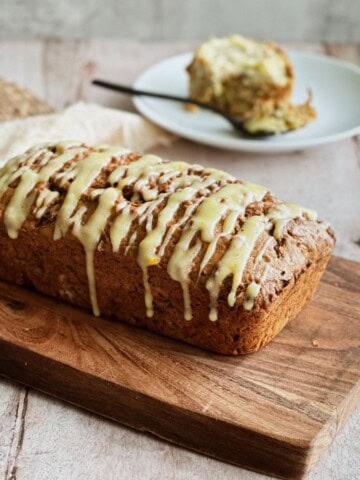 Loaf of zucchini, carrot, and apple bread drizzled with orange cream cheese glaze, resting on a wooden cutting board. A slice is served on a plate in the background with a fork.