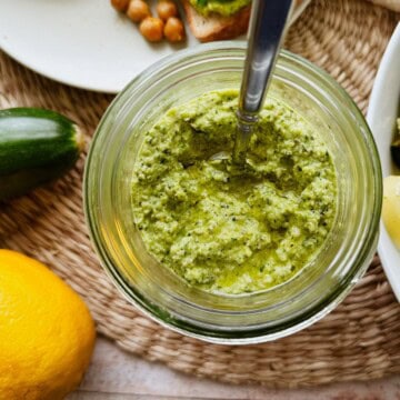 Close-up of an open jar of zucchini pesto with a spoon, surrounded by fresh zucchini and a lemon.