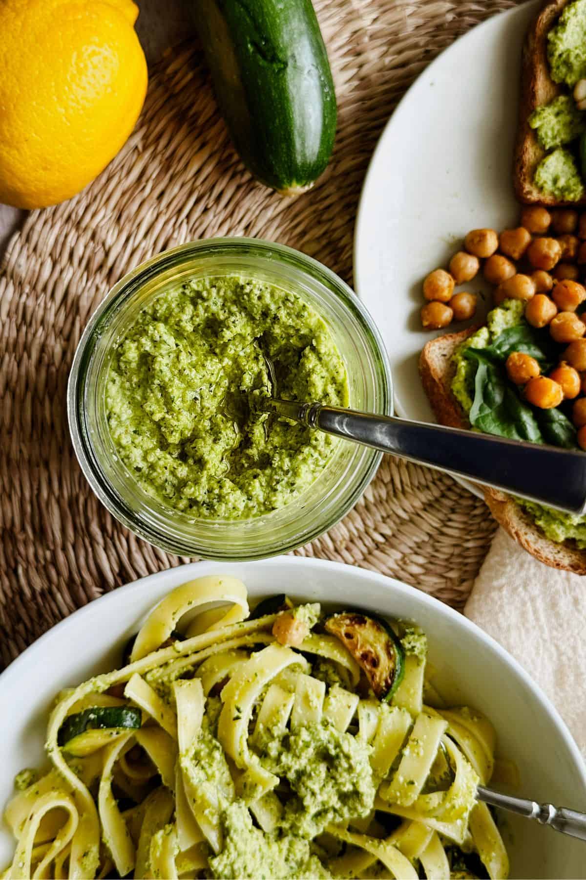Jar of creamy zucchini pesto with a spoon, surrounded by cooked pasta tossed in zucchini pesto and toast topped with zucchini pesto, chickpeas and fresh basil. A fresh lemon and whole zucchini rest nearby on a woven placemat.